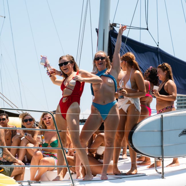 A group of people enjoying a sunny day on a crowded sailboat, some sitting and others standing. The women are wearing bikinis and sunglasses, smiling and holding drinks as a paddleboard is secured to the side of the boat.