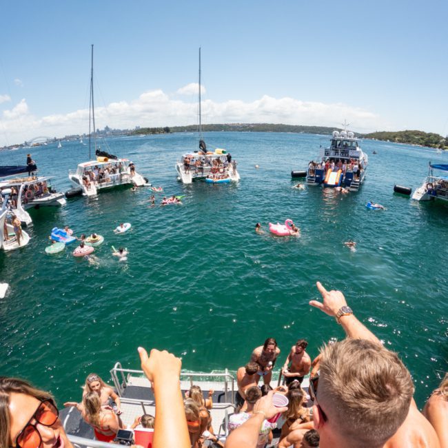 People are swimming and lounging on inflatables between several boats anchored in a sunny, blue water area, enjoying a catamaran party in Sydney.