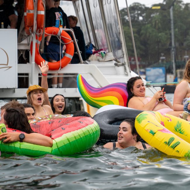 A group of people in swimsuits enjoying a vibrant day in the water with various colorful inflatable floats and a DJ boat hire Sydney near a boat.