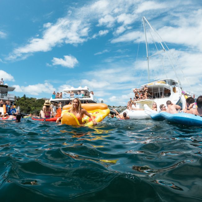 People are enjoying a day on the water with inflatable floats and a luxury yacht hire Sydney in the background under a clear, blue sky.