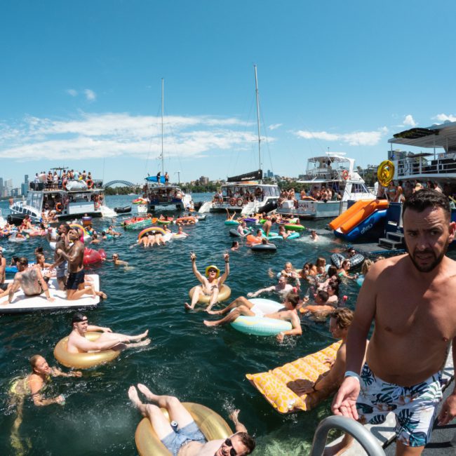 A large gathering of people on boats and inflatables enjoying a sunny day on the water near a city skyline, with some individuals swimming and others lounging on floatation devices. Perfect for those considering a Catamaran party Sydney style.