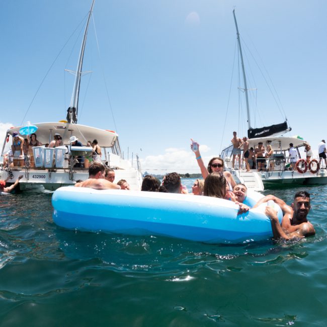A group of people relax in a small inflatable pool floating in the water between two anchored catamarans on a sunny day, enjoying a luxury yacht hire in Sydney.