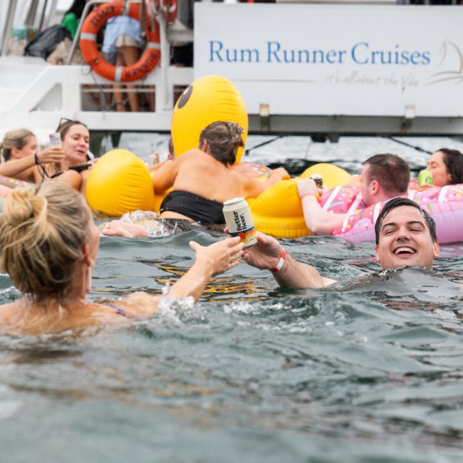 People swimming and floating on inflatable toys near a boat labeled "Rum Runner Cruises," while one person passes a can to another in the water. Perfect for corporate boat events Sydney, it's an idyllic setting combining fun and relaxation.