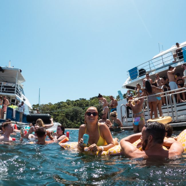 People enjoy a sunny day on the water, swimming and lounging on inflatables, with luxury yachts in the background. The perfect setting for a private yacht charter in Sydney Harbour.