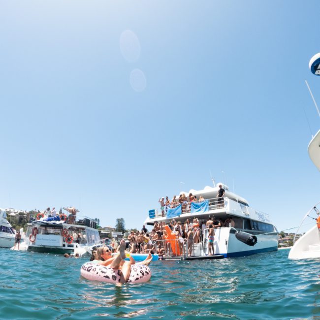 A large group of people enjoying a sunny day on multiple boats anchored close together on a calm body of water. One person is floating in an inflatable raft with others swimming nearby, encapsulating the joy similar to a lively Catamaran party Sydney.