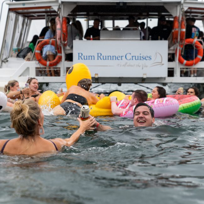 People swimming near a boat named "Rum Runner Cruises," using various inflatable toys. A woman in the foreground holds a drink, while others float and swim around in the water, embodying the fun of a catamaran party on Sydney Harbour.