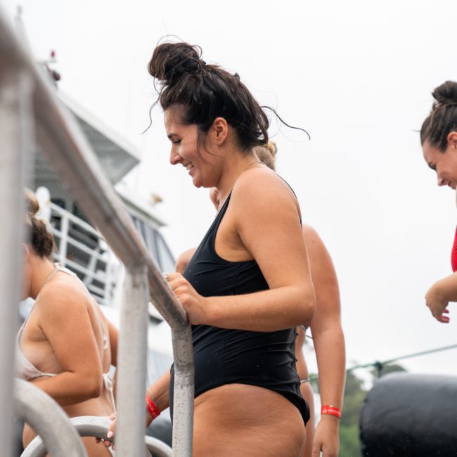 Two women in swimsuits stand on a boat deck, one in black and the other in red, smiling and chatting during a Sydney boat party hire. They are near a metal railing, with another person partially visible in the background.