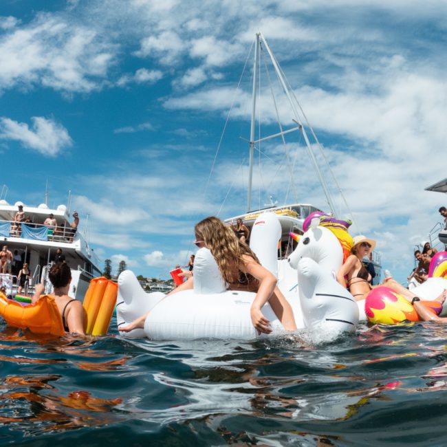 People enjoying a sunny day on the water, floating on various inflatable toys near docked boats, as a private yacht charter sails by in Sydney Harbour.