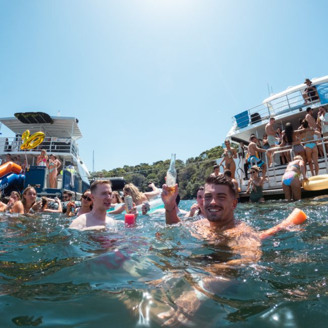 A crowd of people enjoying a sunny day on a lake, some swimming and holding drinks, with two boats anchored nearby, including a lively catamaran party.