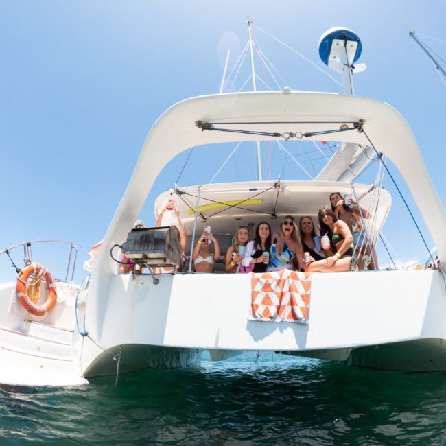 A group of people are gathered on the back of a white catamaran in the ocean on a clear day, enjoying what looks like an epic Catamaran party in Sydney. An orange and white towel hangs on the boat, with a lifebuoy attached to the side.