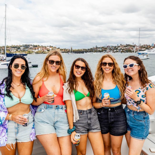 Five women in casual summer attire stand on a boat, smiling and holding beverages. Luxury yachts and a cityscape are visible in the background under a partly cloudy sky.