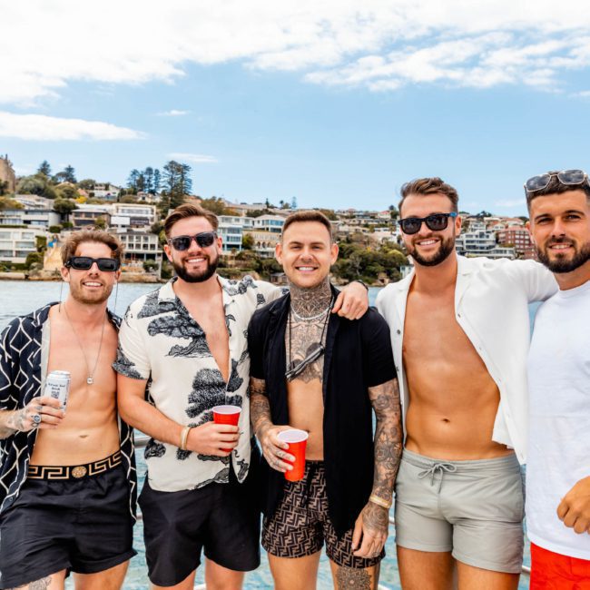 Five men pose together on a boat with water and a coastal town in the background, some holding red cups and one holding a can. They are dressed in casual summer attire, enjoying their private yacht charter on Sydney Harbour.