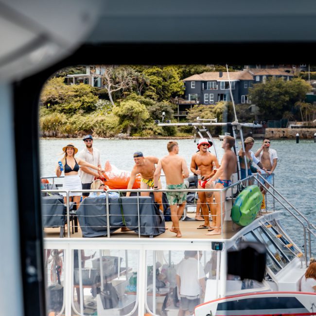 A group of people socializing and enjoying a Sydney boat party hire on a two-level boat near a waterfront, as viewed from within another boat.