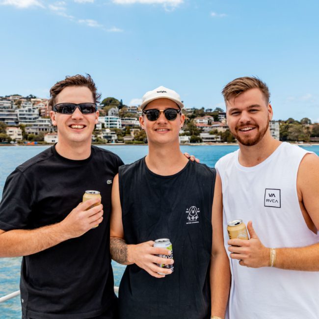 Three men standing on a boat holding drinks, with a scenic view of water and a shoreline with houses in the background, enjoying their Sydney boat party hire.