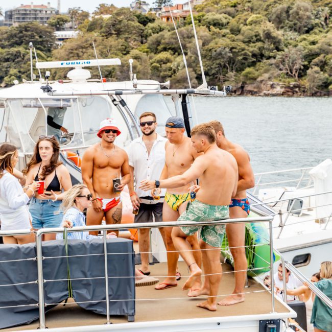 A group of people in swimwear socialize on the deck of a luxury yacht hire Sydney in a scenic waterside location with lush greenery in the background.