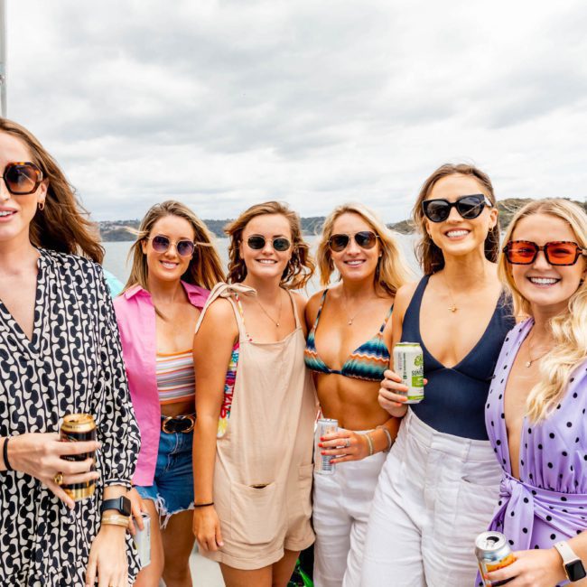 A group of six women stands on a boat, smiling and holding canned drinks. The background shows water and distant land. Dressed for warm weather, they're enjoying their time during a luxury yacht hire in Sydney.