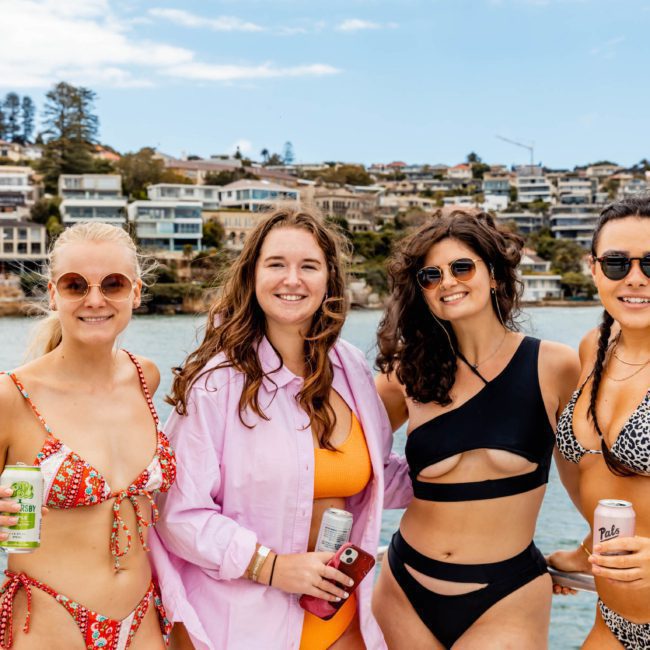 Four women in swimwear pose for a photo on a boat with a scenic view of houses on a hill in the background. They are holding beverage cans and smiling at the camera, enjoying the vibes of a catamaran party in Sydney.