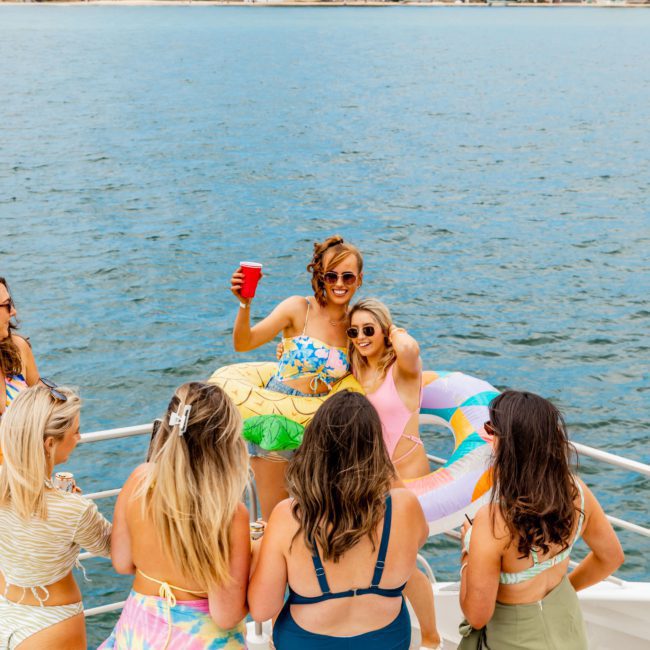 A group of women in swimsuits are gathered on a luxury yacht near the shore. One woman is standing with a drink, wearing sunglasses, with others casually interacting around her.
