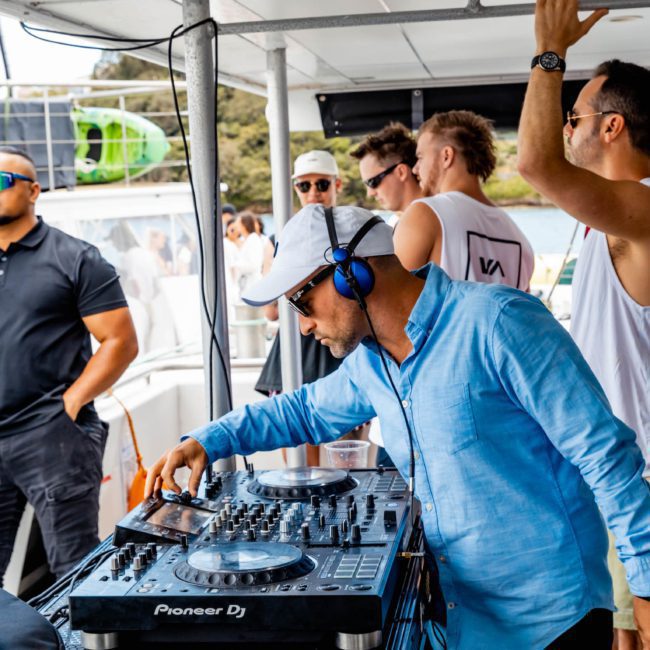 A DJ in a blue shirt and white cap is mixing tracks on a private yacht charter Sydney Harbour while several people in casual attire socialize around him.