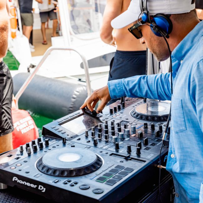 A DJ wearing headphones and a hat operates a DJ controller on a private yacht charter in Sydney Harbour, with people socializing in the background.