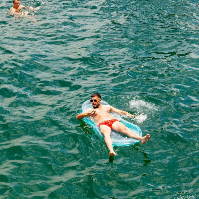 A person reclines on an inflatable float in a body of water, while another person swims nearby, enjoying the serene surroundings of a luxury yacht hire Sydney.