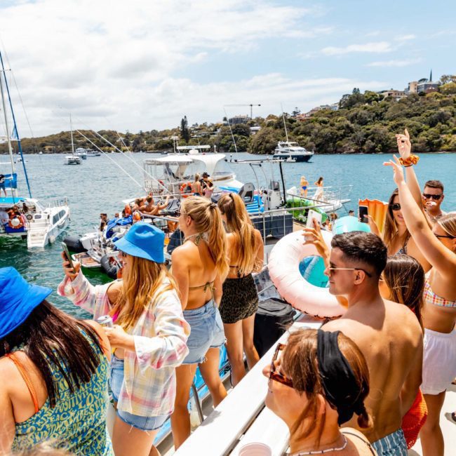 A group of people on a private yacht charter Sydney Harbour is partying, dancing, and taking photos. Several boats are nearby in the water, with additional people swimming and floating on inflatables. Trees and houses are in the background.