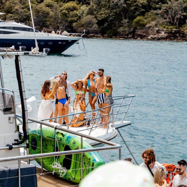 A group of people in swimwear stand at the bow of a yacht, posing for a photo. The yacht is near a forested shoreline, with another boat visible in the background. Perfect for Private yacht charter Sydney Harbour, this scene captures the essence of luxury and relaxation on the water.