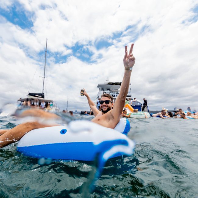 A person relaxes on an inflatable raft in the ocean, holding a drink and flashing a peace sign, with luxury yachts and other people in the background under a partly cloudy sky.