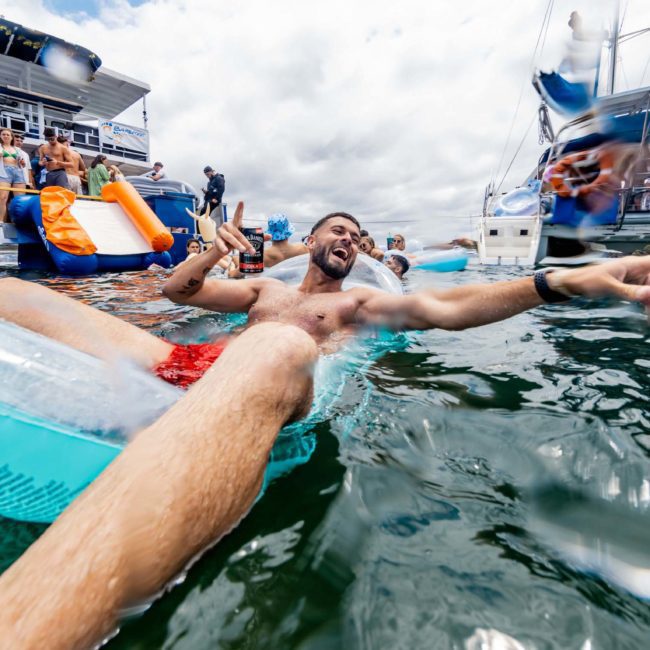 A man lies on an inflatable float in the water, smiling and pointing, while other people are on boats and inflatables behind him. The scene depicts a lively and enjoyable moment on the water, with luxury yacht hire Sydney adding to the atmosphere.