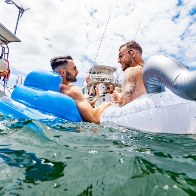 Two men on inflatable floats relax in the water near boats, under a bright, partly cloudy sky during a private yacht charter on Sydney Harbour.