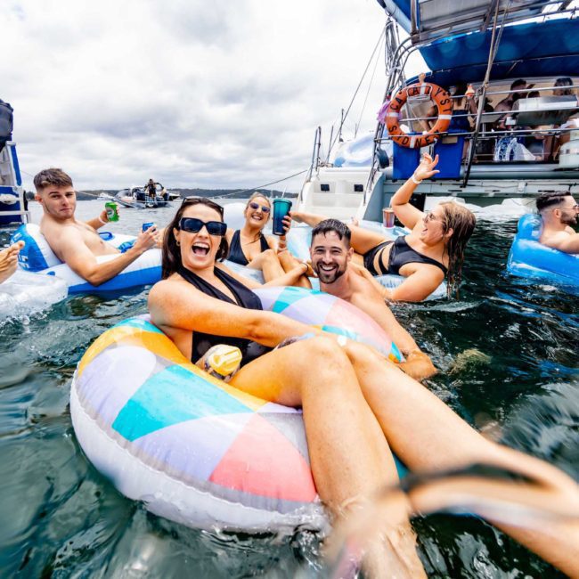 A group of people enjoy a day on the water, many on colorful inflatables and others on boats, socializing and having fun, thanks to Sydney boat party hire.