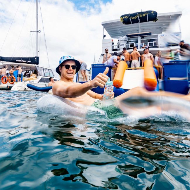 A man in a hat and sunglasses relaxes on an inflatable float with a drink, surrounded by water and yachts, while others gather for a DJ boat hire in Sydney.