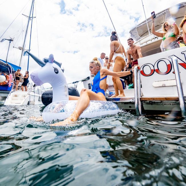 A group of people enjoying a Sydney boat party; some sit on the deck, while others are in the water. A woman is on an inflatable unicorn float. Two boats, perfect for corporate boat events Sydney, are moored close together in the background.