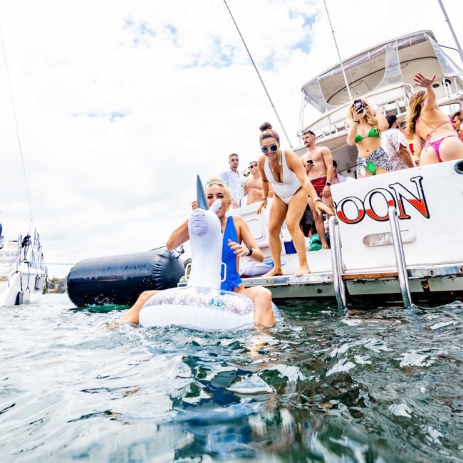 A group of people on the back deck of a boat named "OON," some sitting and some standing, wearing swimwear. One person in the water holds an inflatable object. Perfect for a corporate boat event in Sydney or a private yacht charter on Sydney Harbour.