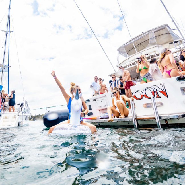 People are gathered on boats and in the water, enjoying a day under the sun. One person is raising their arm triumphantly while sitting on a float in the water. The boat's name "MOON" is visible, perfect for those looking for a Sydney boat party hire or private yacht charter in Sydney Harbour.
