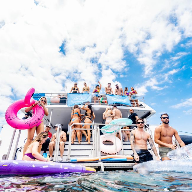 A group of people in swimwear stands on the deck of a boat and in the water, some holding inflatables. The sky is partly cloudy, creating a perfect setting for a private yacht charter Sydney Harbour experience.