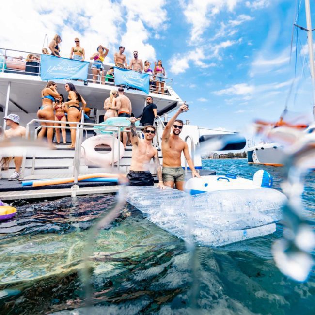 A group of people enjoy a lively party on a boat, with some swimming and lounging on inflatables in the water. Other boats, including DJ boat hire Sydney options, are anchored nearby under a clear sky.