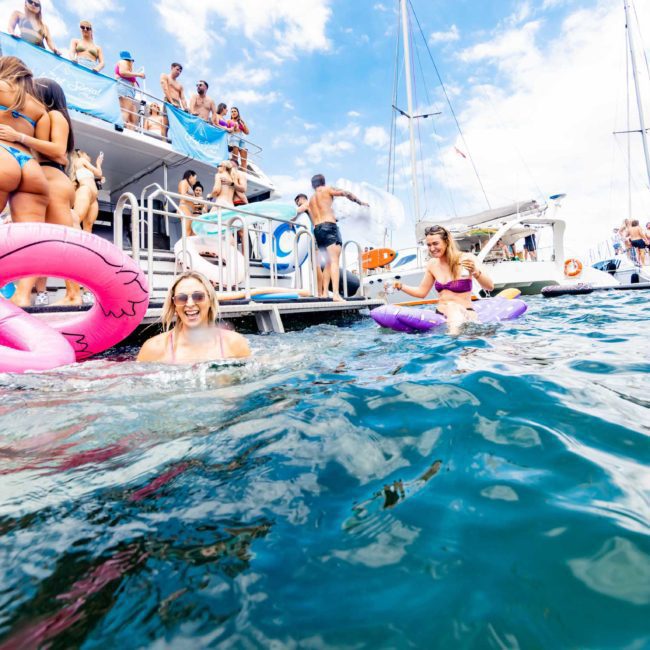 People enjoying a sunny day on a private yacht charter in Sydney Harbour, with some swimming in the water using inflatable floats. Various others are visible on deck and nearby boats, embracing the fun atmosphere of a Sydney boat party hire.