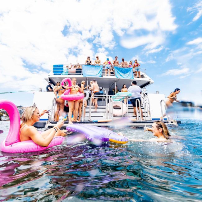 People on a private yacht charter in Sydney Harbour are enjoying their time, with some in the water using inflatable floats shaped like flamingos. Several individuals are gathered on the upper deck of the boat. The sky is partly cloudy.