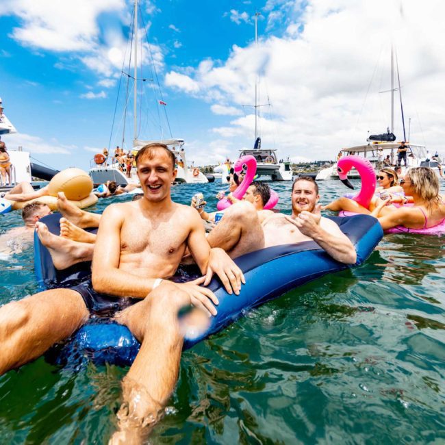 Two men float on an inflatable mattress in the water, surrounded by boats and other people enjoying a sunny day. Among them, several sleek vessels hint at the popularity of corporate boat events in Sydney.