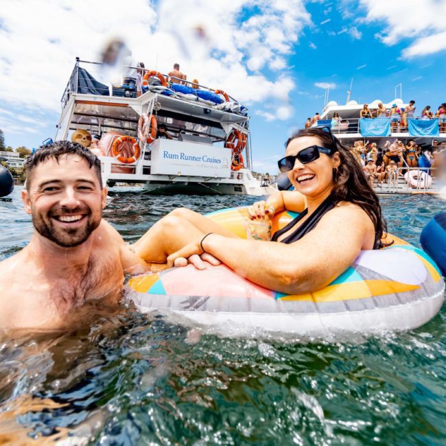 People enjoying a sunny day in the water with boats in the background; a man and a woman in an inflatable float, smiling and relaxed against the backdrop of a luxury yacht hire Sydney.