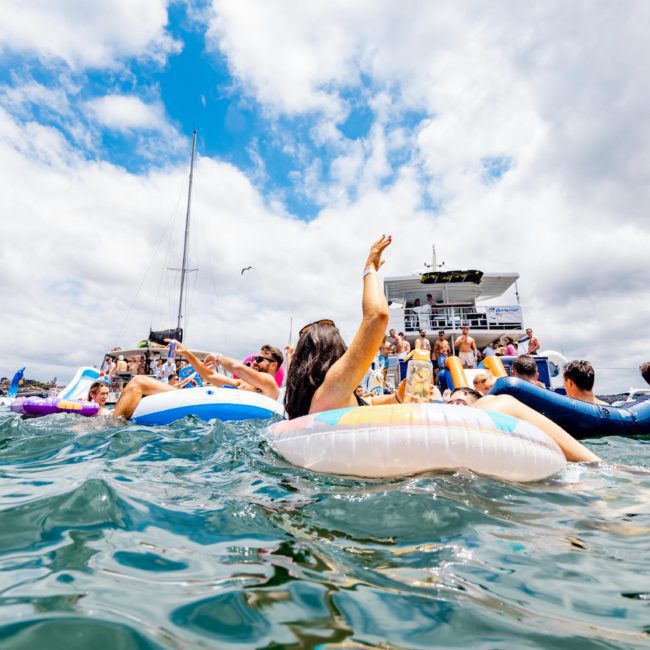 People in swimsuits are floating on inflatable tubes in the water near a DJ boat hire Sydney under a partly cloudy sky.