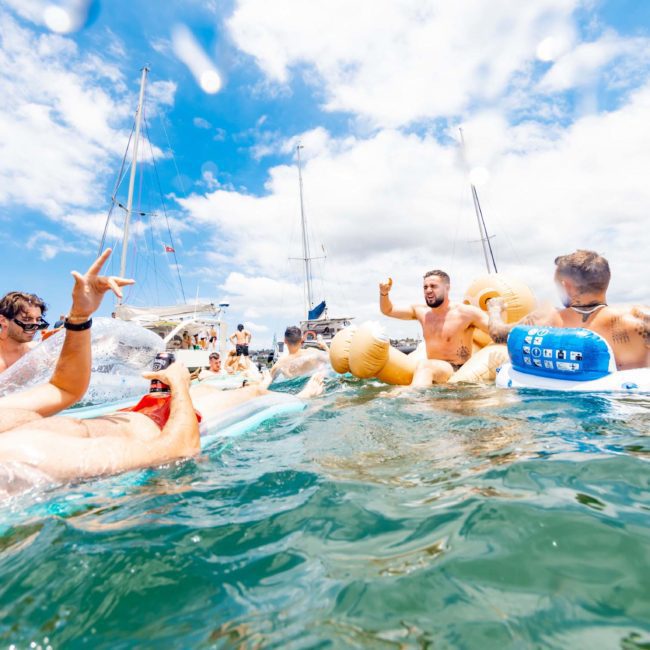 A group of people in swimwear float on inflatable devices in the water near boats under a clear sky, enjoying a DJ boat hire Sydney experience.