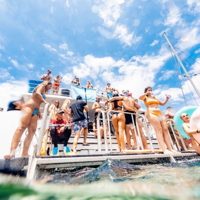 People in swimwear gather on the deck of a boat under a bright sky, with some preparing to enter the water. Droplets on the camera lens give a partial underwater view at this lively Sydney boat party hire.