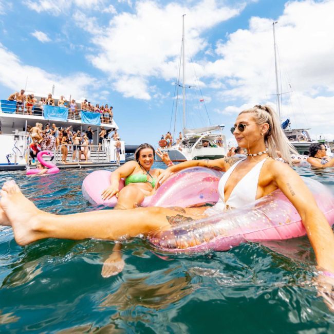 People relaxing on inflatable floats in the water near a boat with onlookers. Some individuals are socializing on a luxury yacht under a partly cloudy sky, enjoying a fantastic experience from Luxury yacht hire Sydney.
