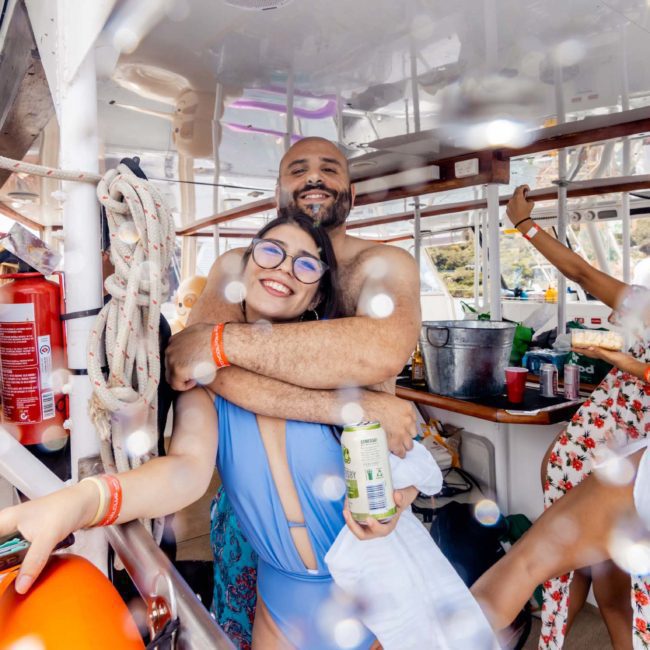 Group of people enjoying a lively Sydney boat party hire, with two individuals in the foreground smiling and holding drinks. The environment is vibrant, featuring life preservers and soaked surfaces on the luxurious yacht.