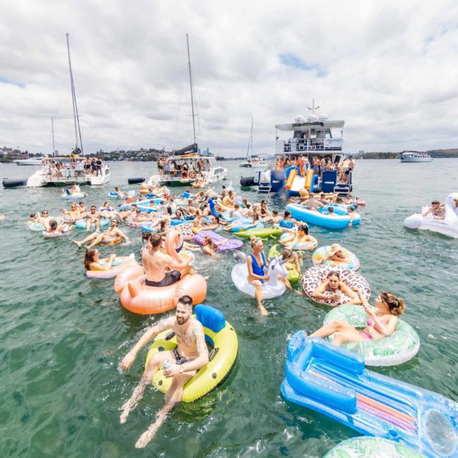 People on inflatable floats and boats enjoy a sunny day on the water. Several yachts and sailboats, including those available for luxury yacht hire Sydney, are docked nearby. The sky is partly cloudy.