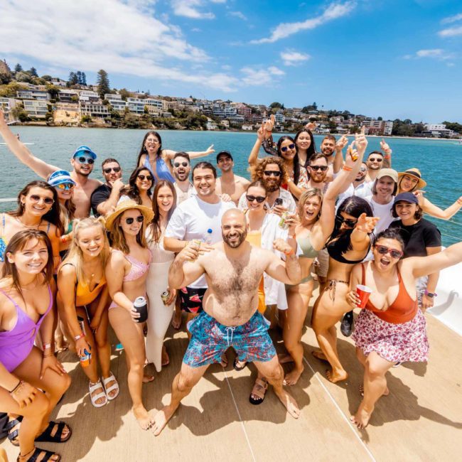 A lively group of people on a catamaran party in Sydney pose for a photo on a sunny day. Some are raising their hands or holding drinks, with houses and trees visible on the shore in the background.