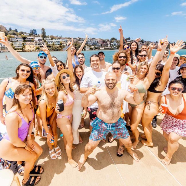A group of people in swimwear are posing and smiling for a photo on the deck of a boat on a sunny day, with waterfront houses in the background, enjoying their DJ boat hire Sydney adventure.