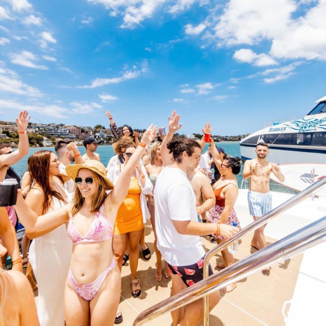 A group of people in swimwear enjoying a sunny day on a boat, with some raising their hands and others mingling. A larger boat with a logo is visible in the background, showcasing the vibrant atmosphere of a DJ boat hire Sydney event.